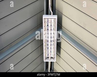 A fluorescent light fixture mounted on gray paneled wall corner, showing rectangular metal housing, reflective grid, wiring, and bright industrial ill Stock Photo