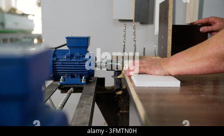 Worker's hands guiding white particleboard into industrial drilling machine with blue electric motor, professional furniture manufacturing Stock Photo