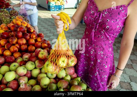 Close-up of a woman in a floral magenta dress shopping at an outdoor market, holding a reusable yellow net bag filled with fresh apples amid piles of Stock Photo