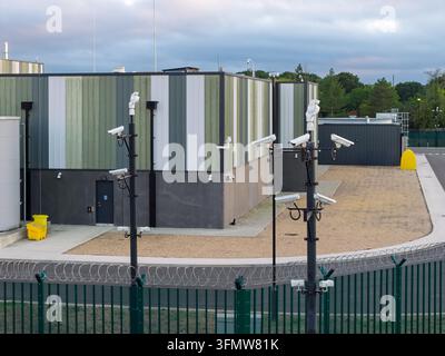 Aerial view of a data center facility featuring security camera poles, perimeter fencing with barbed wire, and gravel grounds in the United Kingdom. Stock Photo