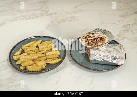Sliced meat wrap halved in foil stacked on a plate, served with salted crinkle-cut fries on marble Stock Photo