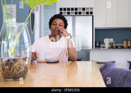 Adult African man sitting at wooden dining table in kitchen holding white mug, smartphone nearby Stock Photo