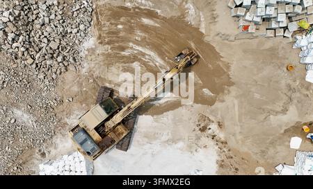 Aerial top-down view of a yellow excavator moving rocks, concrete debris, and soil at active construction site. Heavy equipment performs earthwork. Stock Photo