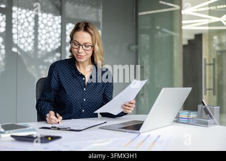 Professional businesswoman wearing glasses examining documents and writing notes while working at a white desk with a laptop, smartphone, and financial graphs in a contemporary office setting Stock Photo