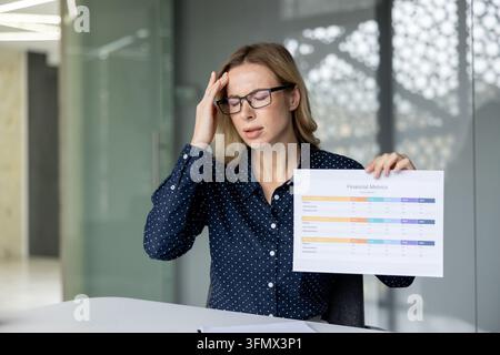 Businesswoman holding a financial metrics report, rubbing her temples in an office, stressed and exhausted by poor performance, overwhelming data and looming deadlines Stock Photo
