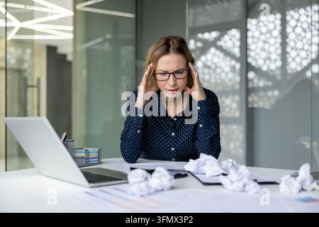 Stressed businesswoman wearing glasses holding temples, experiencing a severe headache and burnout from work, sitting at her office desk with crumpled papers and a laptop Stock Photo