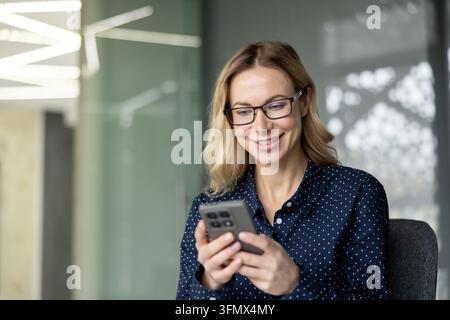 Smiling blonde businesswoman in glasses and a blue polka dot shirt, happily using her smartphone in a modern office, showcasing remote work, digital communication and productivity Stock Photo