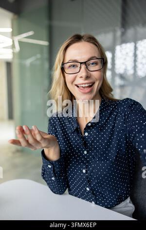 Young blonde businesswoman wearing eyeglasses smiling and gesturing while participating in an online video call or virtual meeting from a modern office workspace Stock Photo