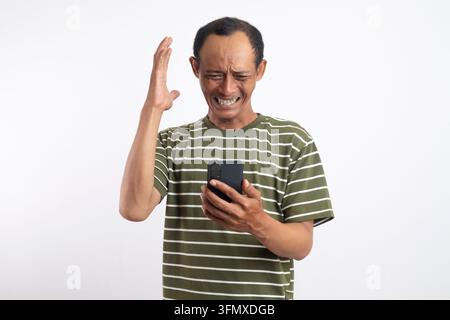 Young Asian man wearing green shirt looking at his smartphone with a stressful expression, isolated by white background. Stock Photo