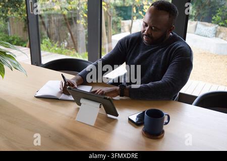 African man wearing dark sweater sitting at home at wooden table writing in notebook using tablet Stock Photo