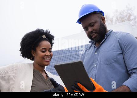 African American team inspecting tablet at rooftop panels, tech wearing blue hardhat, orange gloves Stock Photo