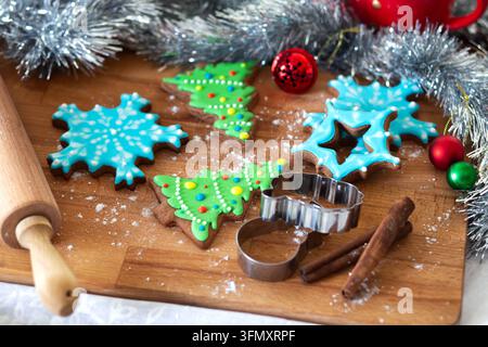 Cookies shaped like trees, snowflakes, and stars are decorated with icing and placed on a wooden countertop. Baking tools and decorations are present. Stock Photo