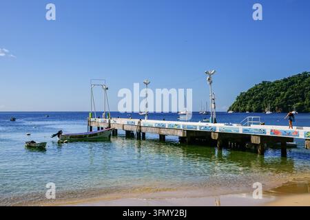 The Charlotteville fishing pier, its railing painted with murals of tropical fish, stretches into the clear water of Man of War Bay, with the fishing boat "Humble Soul" moored alongside and sailboats anchored in the bay, at Charlotteville, Tobago, Trinidad and Tobago. Stock Photo