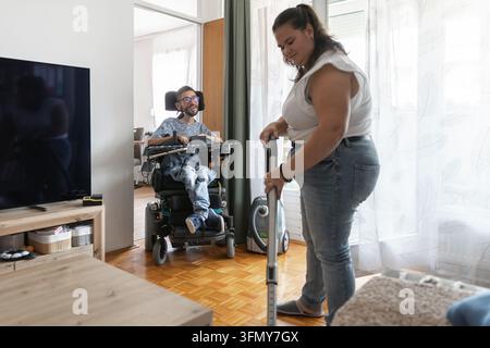A woman vacuuming floor the floor with a man in a wheelchair in their home. They are engaged in daily living activities together. Stock Photo