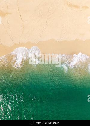 Aerial vertical landscape directly above white waves gently breaking on soft, golden sand scattered with footprints in a tropical paradise or travel c Stock Photo