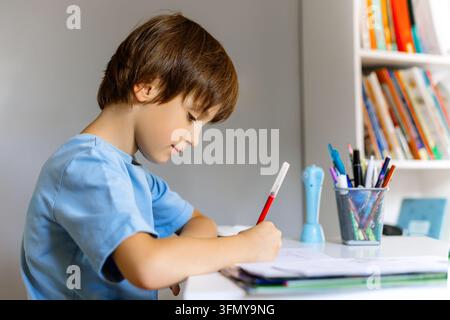 A focused schoolboy in a blue t-shirt sits at a desk, writing with a red marker. Smart kid doing homework at home, school education concept, selective Stock Photo