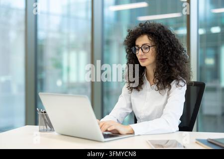Young businesswoman with curly hair and glasses focusing on her work, typing on a silver laptop at a clean white desk in a contemporary office setting, representing productivity and technology Stock Photo