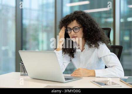Stressed young business woman with curly hair and glasses holding her head, experiencing a headache and burnout while working on a laptop in a modern office Stock Photo