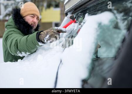 Bearded man in winter clothing removes heavy snow and ice from a car windshield with a red scraper outdoors, preparing his vehicle for safe travel in Stock Photo