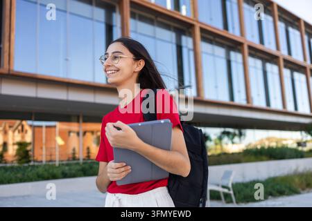 Smiling young woman wearing glasses and a backpack holds a tablet while walking outside a modern university building, portraying student life, educati Stock Photo