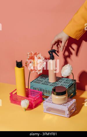 Colorful cosmetic containers, jars, and bottles stand on plastic crate pedestals with flowers against a yellow and pink background as a woman’s hand r Stock Photo