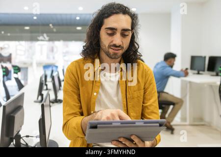 Young man uses a tablet at a shared desk in a bright modern coworking office, while another professional works at a computer workstation in the backgr Stock Photo