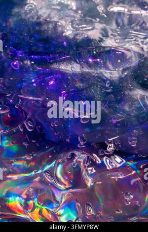 Close-up abstract texture of melting ice cubes and trapped air bubbles in water, illuminated by vivid purple, blue, and rainbow iridescent reflections Stock Photo