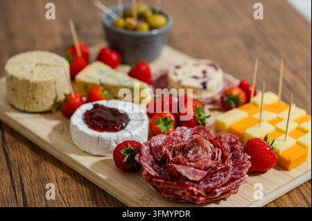 Assorted cheeses, cured meats, fresh strawberries, and green olives arranged on a wooden cutting board, creating an inviting charcuterie spread for sh Stock Photo