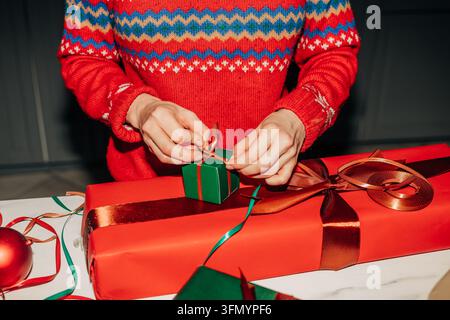 Close-up of a woman in a festive red sweater wrapping Christmas gifts with green and gold ribbon on a table, creating a warm seasonal holiday preparat Stock Photo
