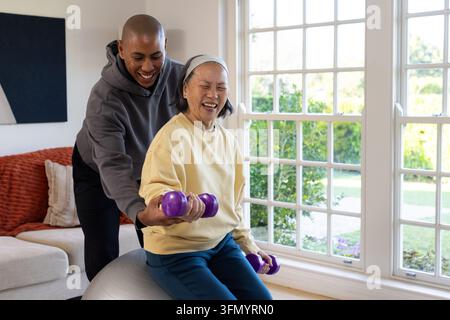 Diverse trainer guiding senior Asian woman exercising at home on silver ball holding purple weights Stock Photo