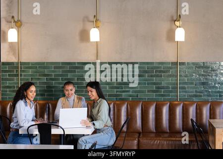 Diverse female coworkers collaborating around white laptop at cafe with green subway tiles. Collaboration, teamwork, professionals, coworking, minimal Stock Photo