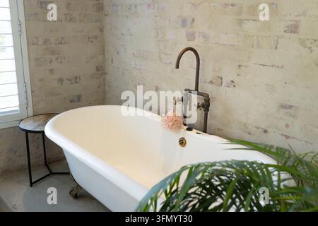 White clawfoot bathtub is sitting against painted brick wall with vintage faucet and pink pouf Stock Photo