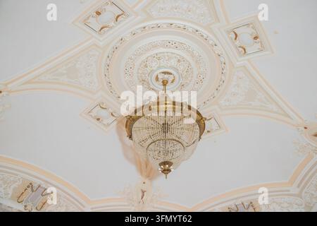 Luxury interior lighting fixture with detailed plaster ornament on white ceiling.Bottom view of grand vintage crystal chandelier hanging from Stock Photo