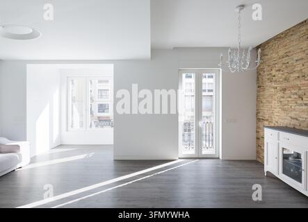Bright luxury house interior with minimalist design, gray flooring, white walls, exposed brick wall, chandelier and abundant sunlight streaming throug Stock Photo