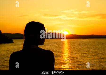 Dark silhouette of a woman gazing across a tranquil lake as the sun sets behind distant hills, casting a glowing golden reflection across the water. Stock Photo