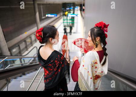 Two women wearing elegant flamenco dresses and red flower hair accessories hold decorative fans while descending an escalator inside a modern metro st Stock Photo