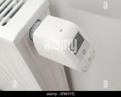 White smart thermostatic radiator valve showing digital display mounted on ribbed panel radiator against light wall, close-up of programmable home hea Stock Photo