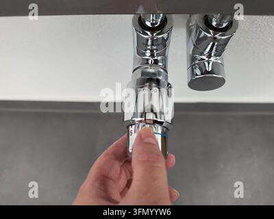 Person turning thermostatic radiator valve on chrome heating pipes mounted on grey wall, close-up view of manual temperature adjustment in home interi Stock Photo