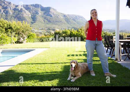Woman wearing red blouse and jeans standing smiling on sunlit lawn with golden retriever Stock Photo