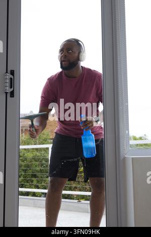 African man cleaning sliding glass door on balcony using squeegee, spray bottle, headphones Stock Photo