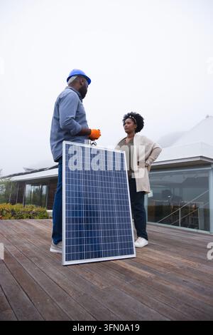 African technician showing owner, holding wires on rooftop near solar panel, in helmet and gloves. Photovoltaic, array, outdoor, terrace, deck, glass, Stock Photo