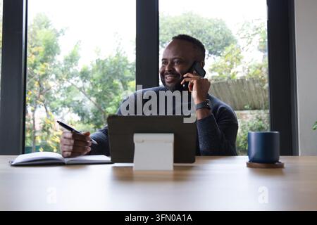 African American man smiling talking on phone holding pen over notes at table with tablet stand Stock Photo