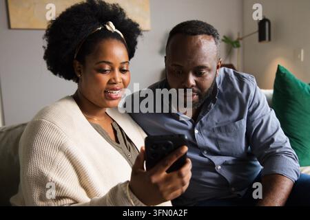 Couple sitting closely on light-colored sofa sharing black smartphone, woman wearing cream cardigan Stock Photo