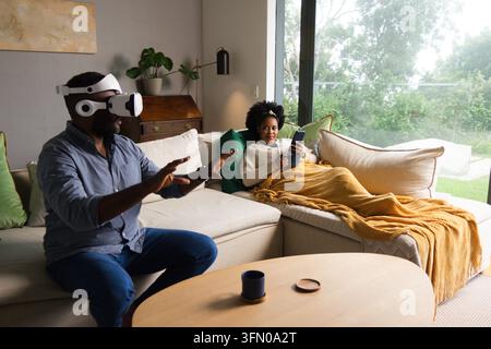 African American couple relaxing at home, man wearing VR, woman scrolling phone under mustard throw Stock Photo