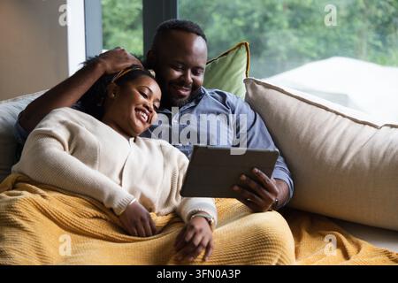 African couple sitting on sofa, snuggling under mustard-yellow knit blanket and holding tablet Stock Photo