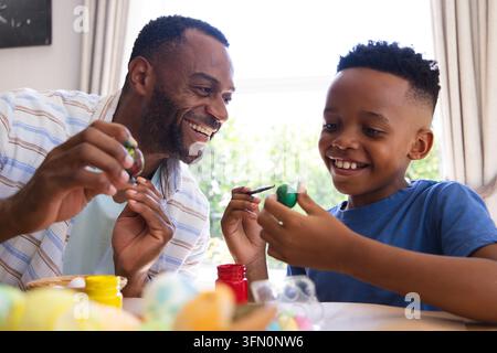 African American father and child painting eggs with paint jars and brushes at table by window Stock Photo