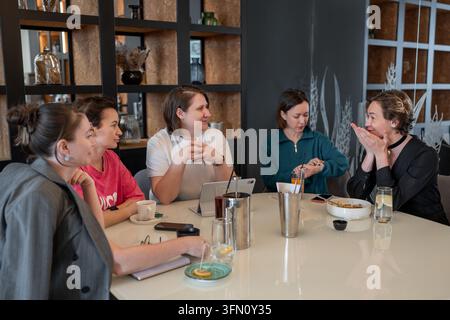 Group of female colleagues enjoying lunch and conversation around round table in modern restaurant, warm lighting, glass partition with frosted Stock Photo
