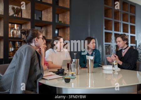 Group of female colleagues enjoying lunch and conversation around round table in modern restaurant, warm lighting, glass partition with frosted Stock Photo