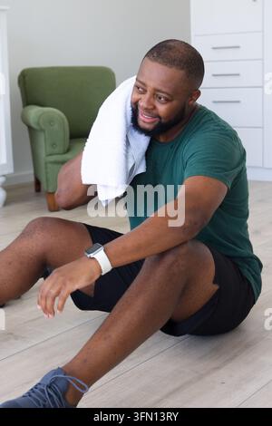 Adult African American man sitting on floor at home, wiping neck with towel, wearing green T-shirt Stock Photo