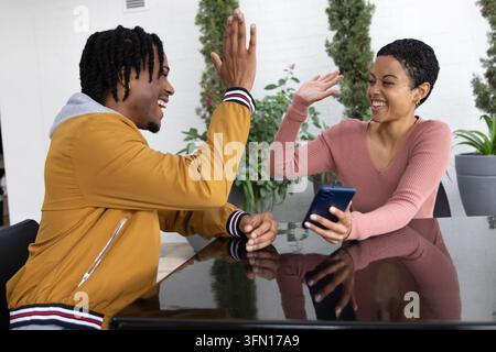 African American couple high-fiving at patio, phone on table, plants behind, wearing casual clothes. Outdoor, courtyard, greenery, planters, glossy, smartphone, bright Stock Photo